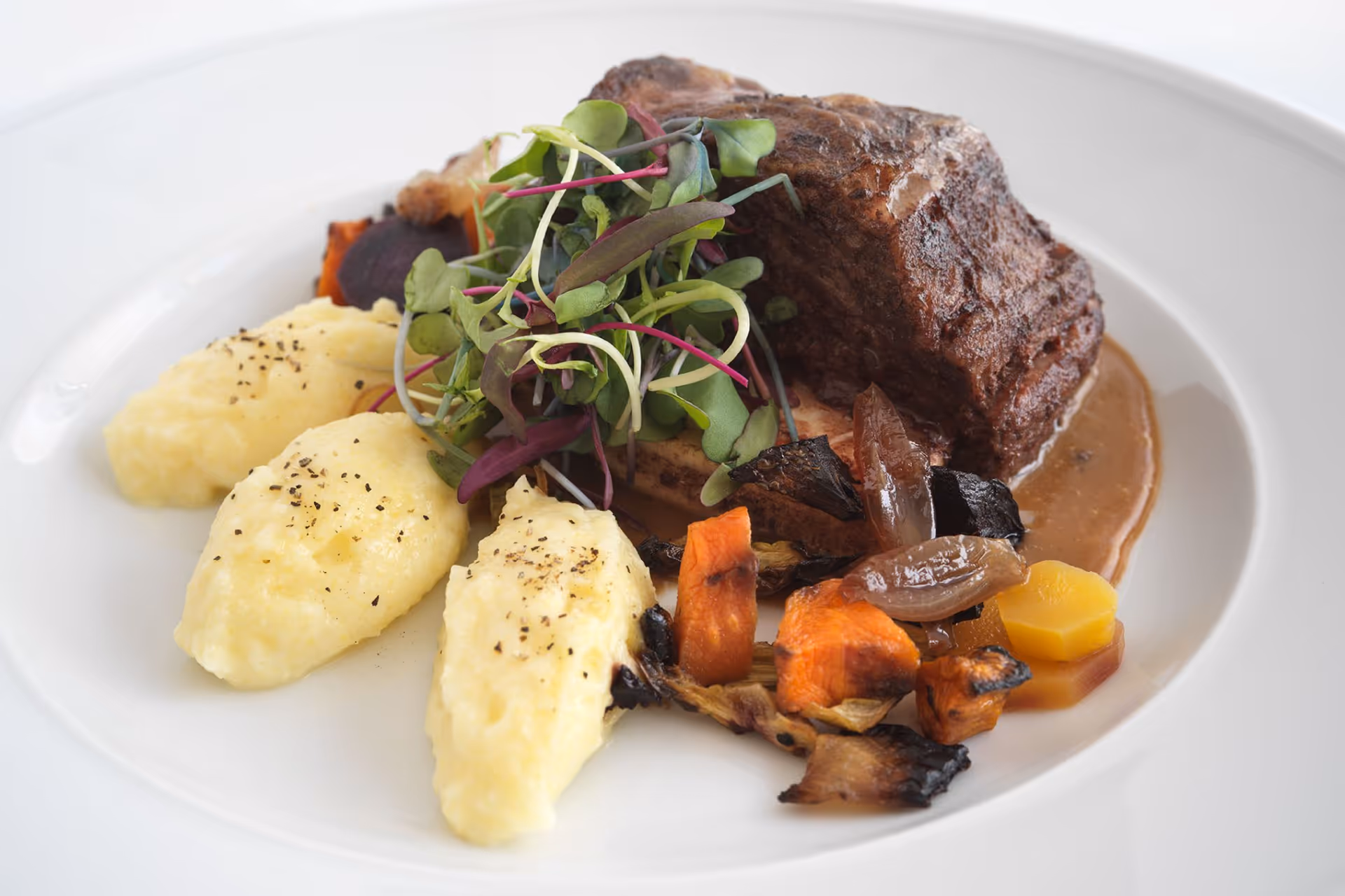 A plated gourmet meal featuring a piece of braised beef with brown sauce, mashed potatoes sprinkled with black pepper, roasted vegetables, and a garnish of fresh microgreens on a white plate.