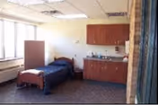 A small room with a single bed covered with a dark blue blanket, a wooden headboard and footboard, a window with blinds on the left side, and a kitchenette area with cabinets, a sink, and countertop on the right side.