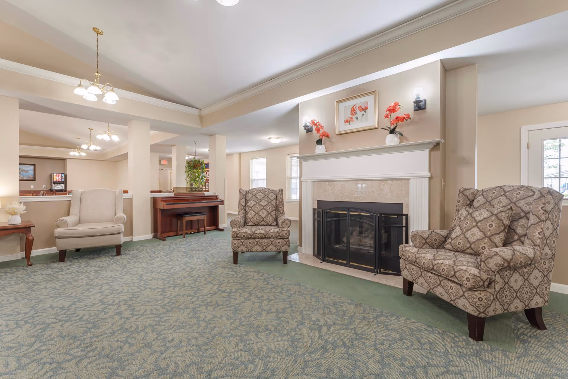 A spacious living room area with patterned carpet, three upholstered armchairs, a fireplace with a white mantel decorated with two vases of red flowers and a framed floral picture above it. There is a piano against the far wall and multiple ceiling light fixtures providing warm lighting.