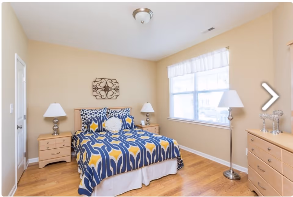 A bedroom with a bed covered in a blue, yellow, and white patterned comforter and matching pillows. There are two wooden nightstands with lamps on either side of the bed, a wooden dresser with decorative items, a floor lamp, and a large window with white curtains letting in natural light. The walls are beige and the floor is wooden.