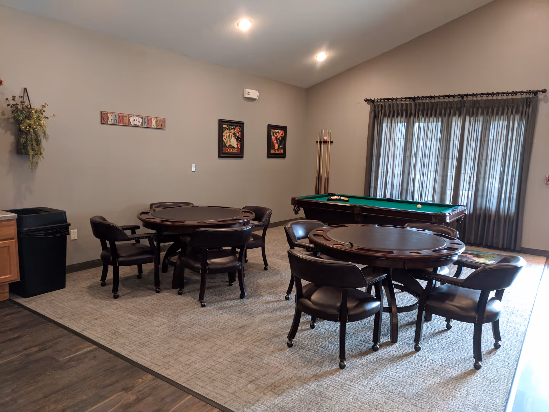 A game room with two round card tables each surrounded by four dark wooden chairs with leather seats. In the background, there is a pool table with a green felt top and pool cues standing in a corner. The room has beige walls with framed pictures and a sign that reads 'GAME ROOM'. A window with sheer curtains allows natural light to enter the room.