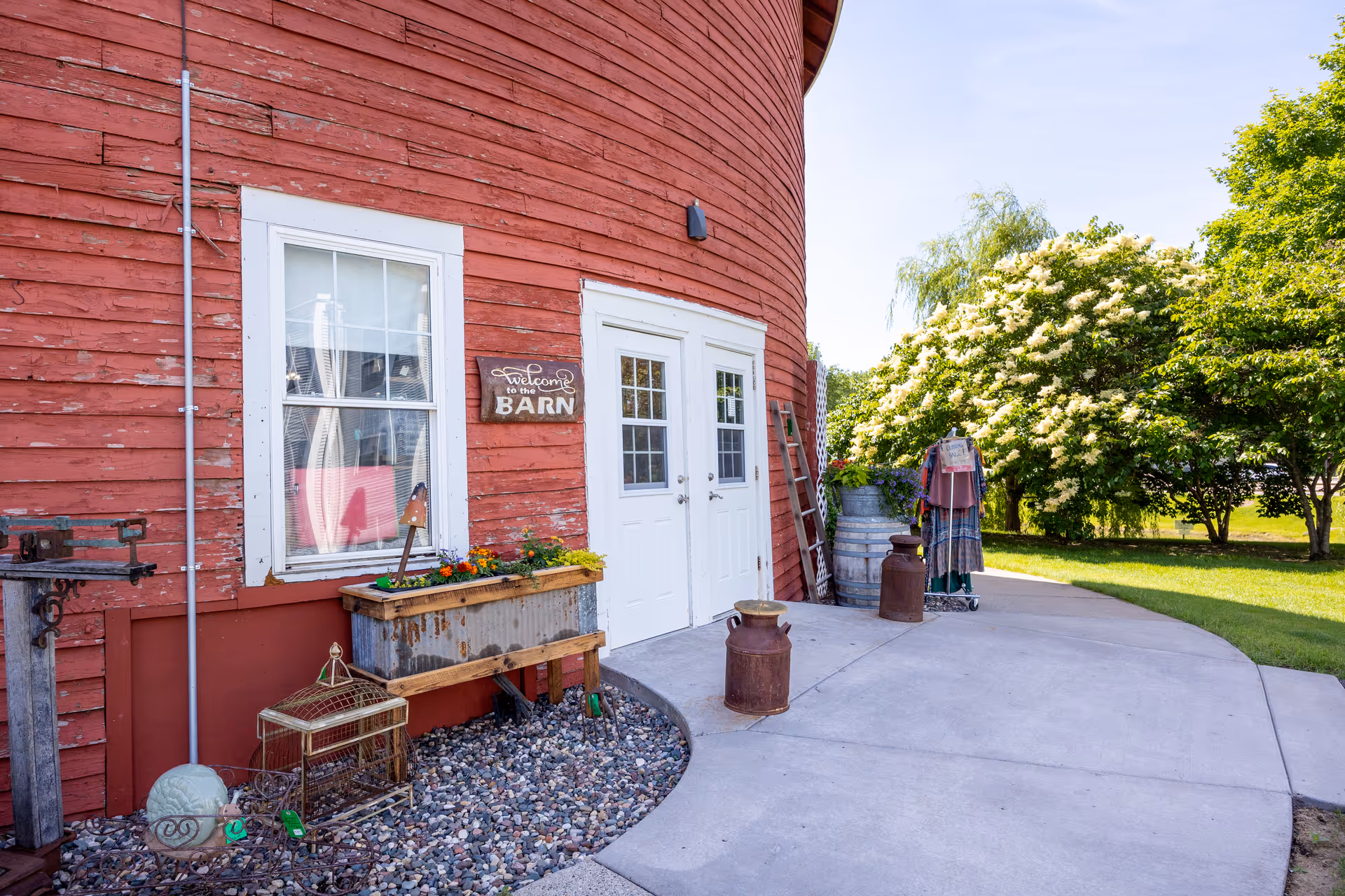Exterior view of a red barn-style building with white double doors and a window. There is a wooden sign that says 'Welcome to the Barn' next to the door. Various decorative items including flower boxes, old milk cans, a ladder, and a clothing rack are placed outside. Green trees and a clear sky are visible in the background.