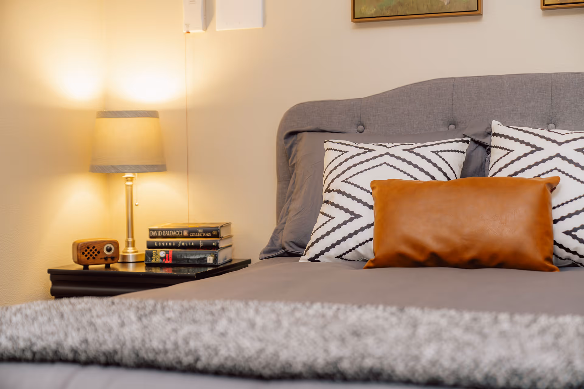 A neatly made bed with a gray upholstered headboard, two white pillows with black zigzag patterns, and a brown leather rectangular pillow. A gray blanket is folded at the foot of the bed. To the left, there is a dark wooden nightstand with a small vintage-style radio, a stack of books, and a lit table lamp with a beige shade. The wall behind the bed is light-colored with a framed picture partially visible above the headboard.
