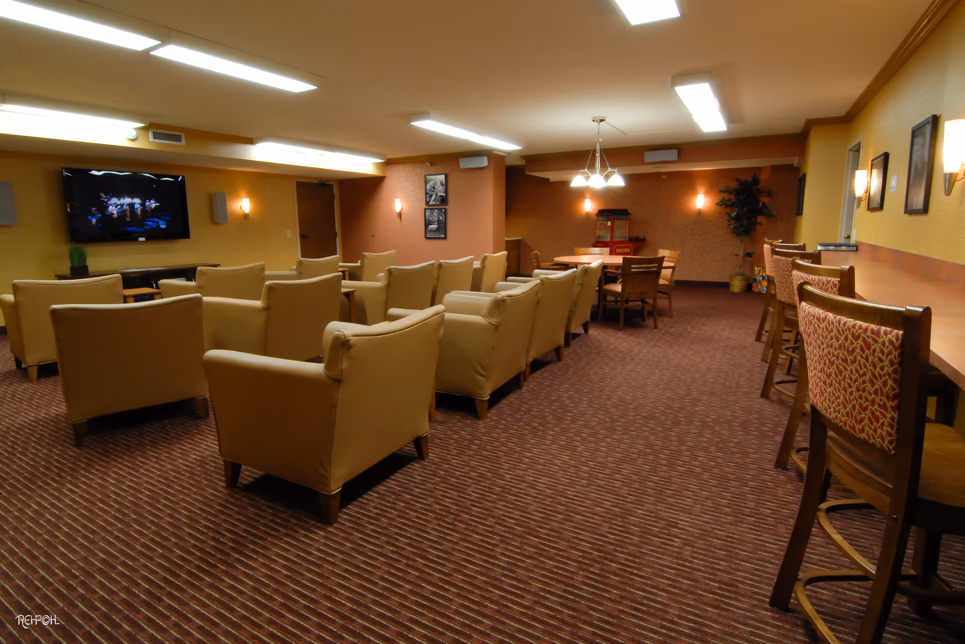 A cozy common area with rows of beige armchairs facing a wall-mounted television. To the right, there is a counter with high chairs and a round table with chairs in the background. The room has warm lighting, carpeted floor, and framed pictures on the walls.