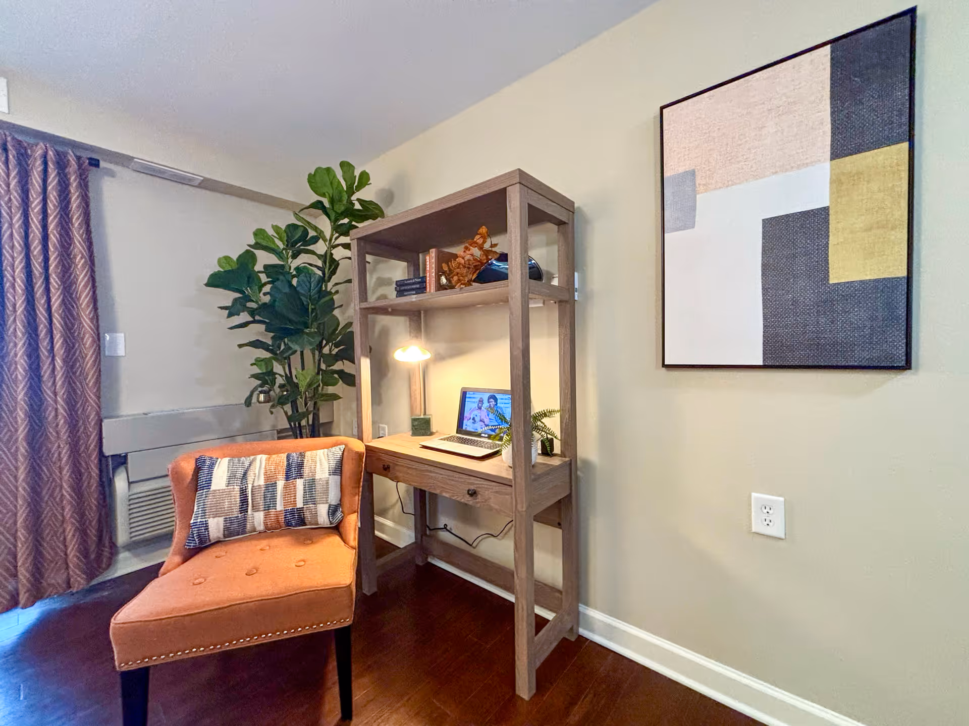 Cozy living-room corner with an upholstered chair, a wooden desk with shelving holding a laptop and lamp, a potted plant, and abstract wall art.