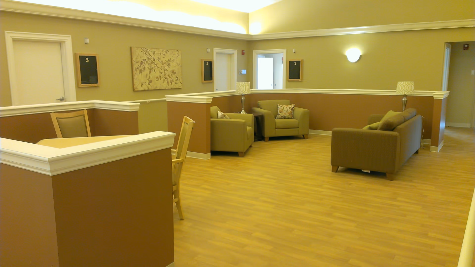 A well-lit interior common area with light brown walls and wood flooring. The space includes a small seating area with two armchairs and a sofa, each with cushions, and two table lamps on side tables. There is a small table with chairs near a half-wall partition. Three doors are visible in the background, each labeled with numbers 1, 2, and 3. A decorative wall art piece hangs between the doors.