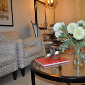 A cozy living room area featuring a patterned armchair, a glass coffee table with a red book and a decorative metallic sculpture, and a vase with white flowers. In the background, there is a dining table with chairs and a chandelier hanging above.