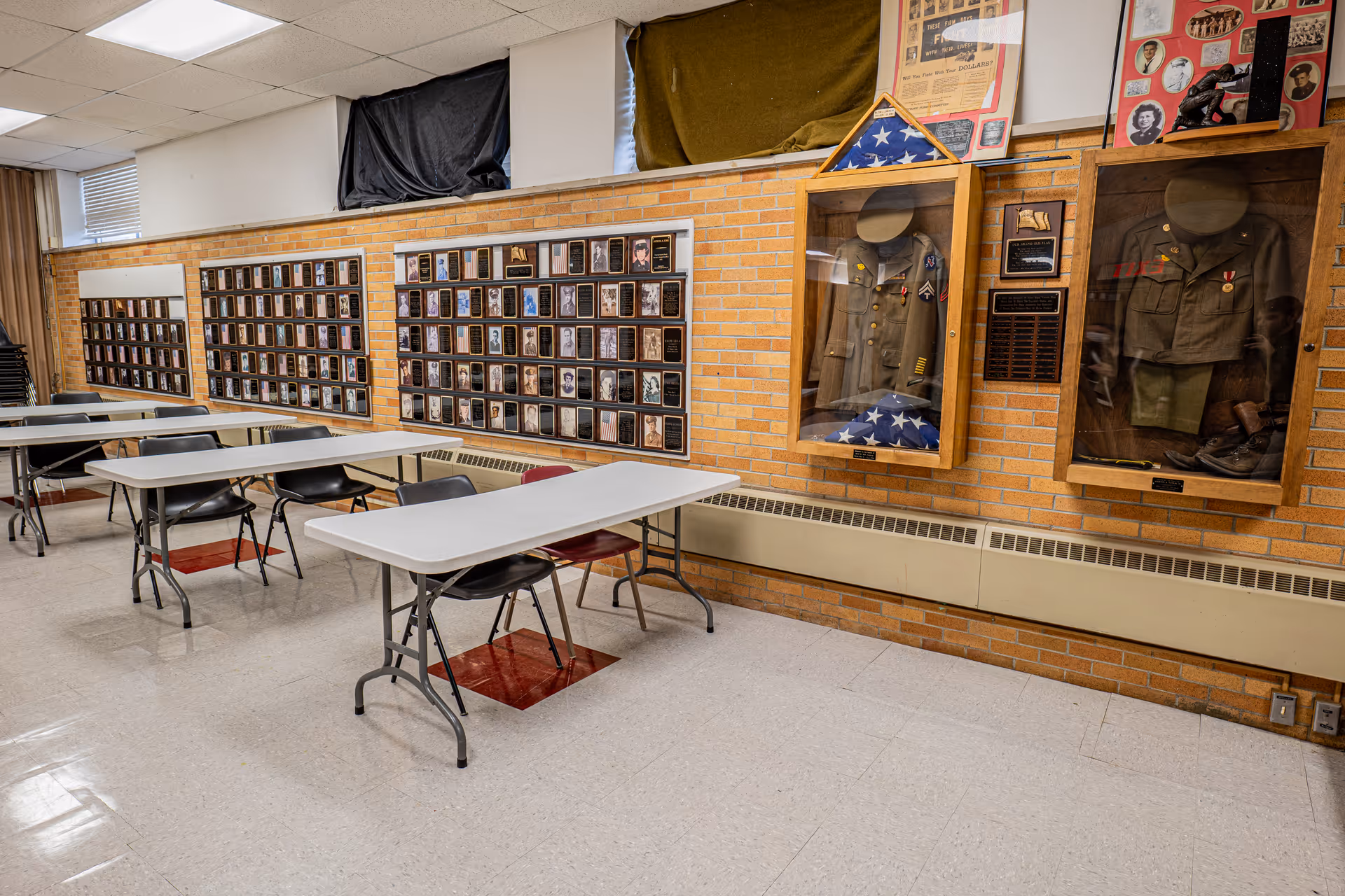 Interior room with folding tables and chairs arranged in rows. The walls are decorated with numerous framed photographs and two display cases containing military uniforms and folded American flags. The room has a tiled floor and a brick wall background.