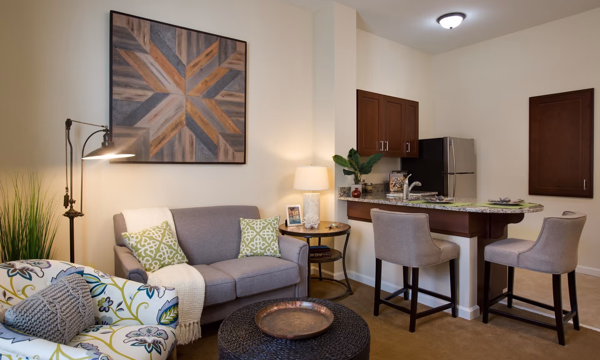 A cozy living area with a gray loveseat adorned with two green patterned pillows and a white throw blanket. Next to it is a round side table with a lamp and decorative items. A floral-patterned armchair with a gray knitted pillow is partially visible. The room includes a kitchen area with a granite countertop island, two beige upholstered bar stools, wooden cabinets, and a stainless steel refrigerator. A large geometric wooden wall art hangs above the loveseat.