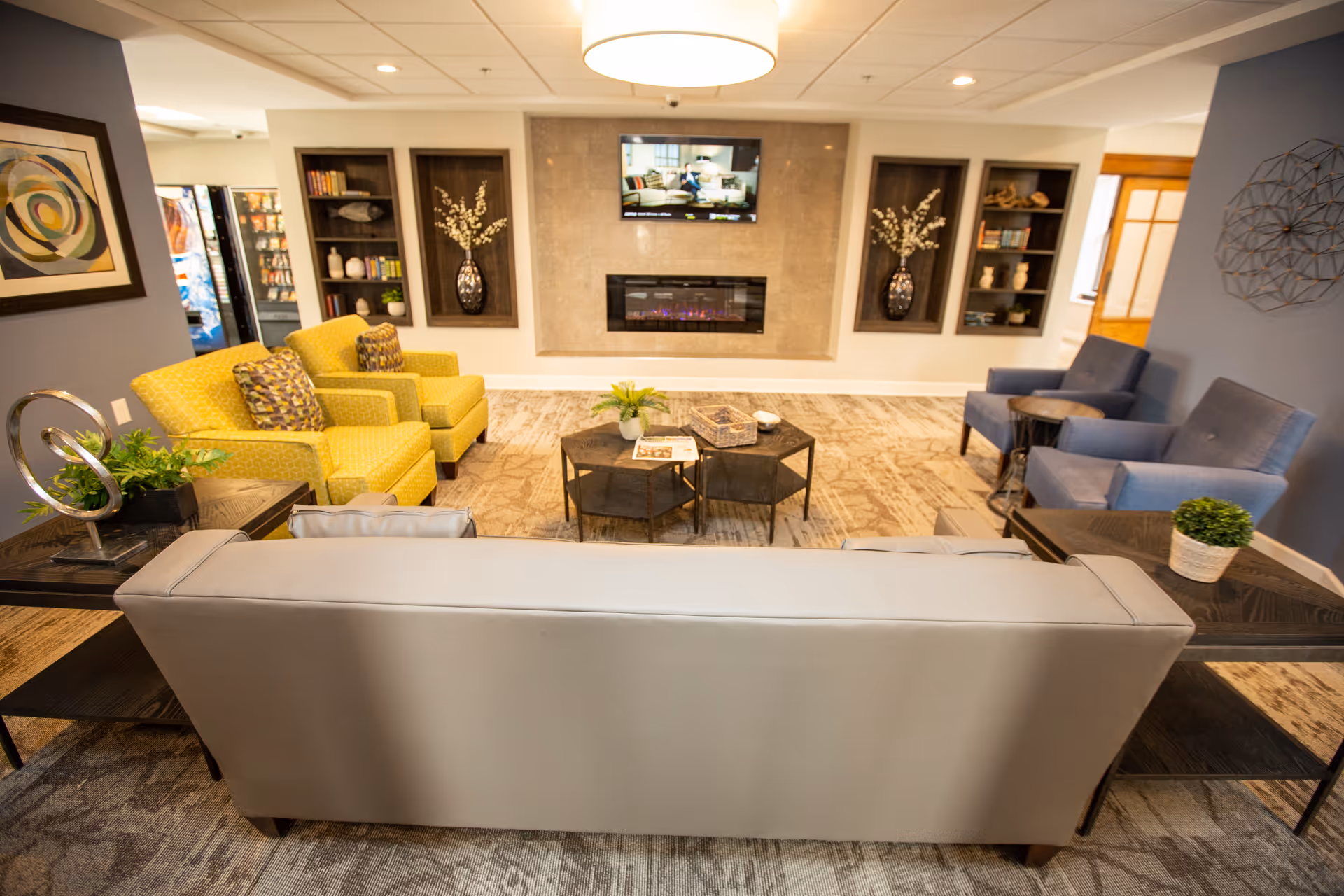 A cozy senior living facility lounge area with a gray sofa in the foreground, two yellow armchairs on the left, and two blue armchairs on the right. A modern electric fireplace is mounted on the far wall beneath a flat-screen TV. Built-in shelves with decorative items and plants flank the fireplace. The room has a warm, inviting atmosphere with soft lighting and carpeted flooring.