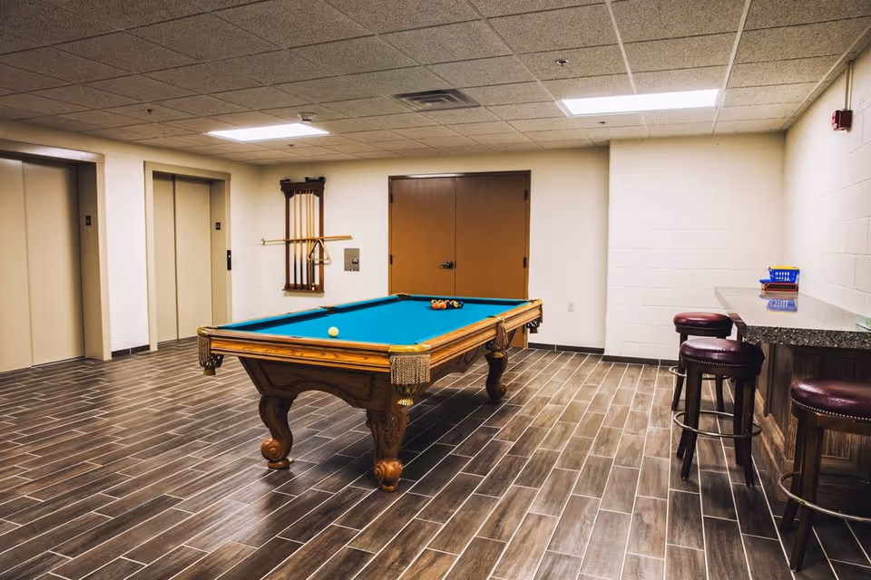 A recreational room with a pool table in the center, a rack with pool cues mounted on the wall, two elevators on the left side, a double door in the background, and a counter with three bar stools on the right side.