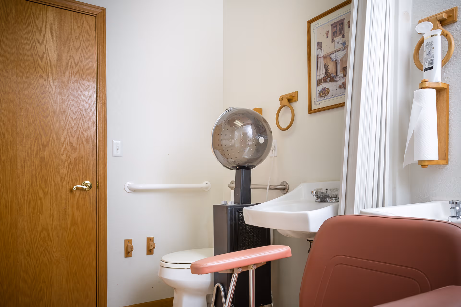 A bathroom with a wooden door, a white toilet, a white sink, a wall-mounted paper towel holder with a roll of paper towels, a framed picture on the wall, and a vintage hair dryer chair with a pink seat.
