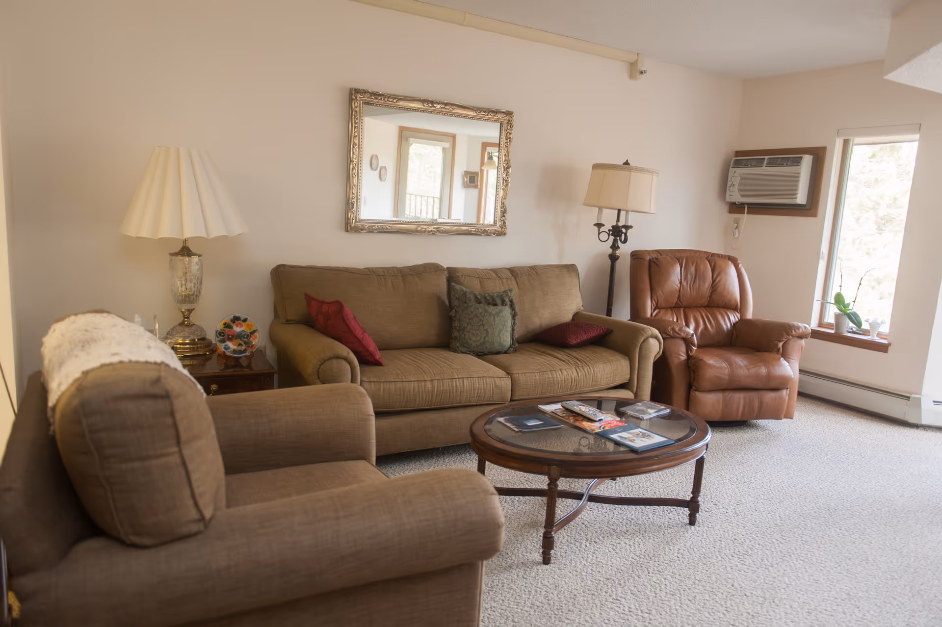 A cozy living room with a brown fabric sofa, matching armchair, and a brown leather recliner. There is a round wooden coffee table with magazines on it in the center. A decorative table lamp is on a side table next to the sofa, and a floor lamp is beside the recliner. A large ornate mirror hangs on the wall above the sofa, and a window air conditioning unit is installed in the wall near a window with a small plant on the windowsill.