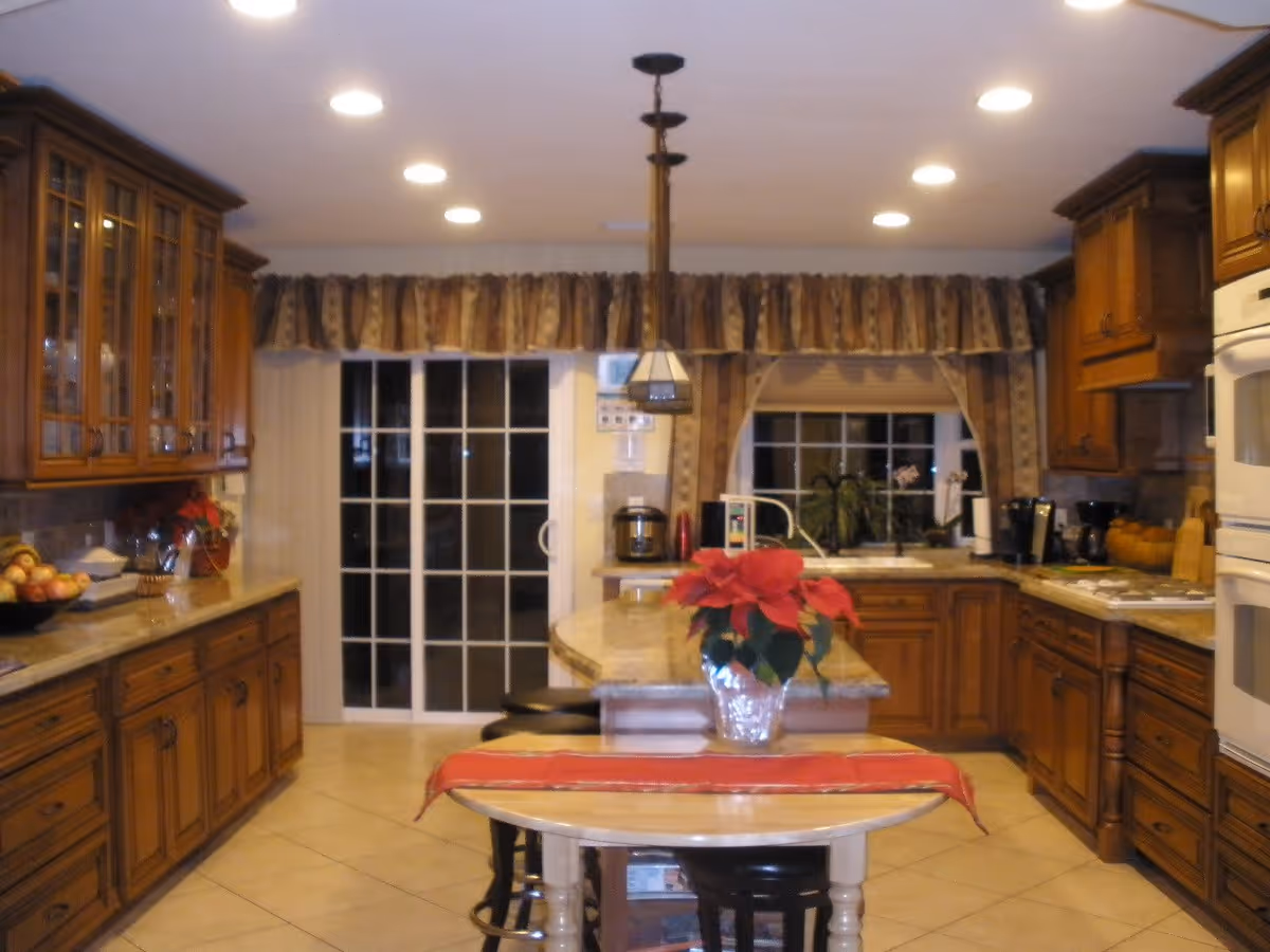 A spacious kitchen with wooden cabinets and drawers, a central island with a red poinsettia plant on top, and a small table with a red runner in the foreground. The kitchen has tiled floors, recessed ceiling lights, a sliding glass door with curtains, and various kitchen appliances including a coffee maker and microwave.