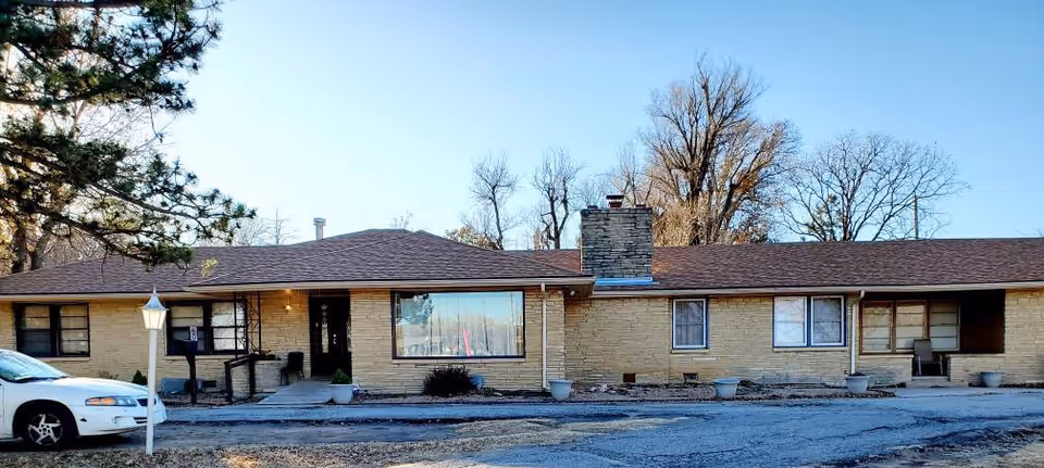 Single-story brick building with a brown shingled roof, a large front window, a chimney, and a small porch area. There is a white car parked on the left side and a lamp post near the entrance. Leafless trees are visible in the background under a clear blue sky.