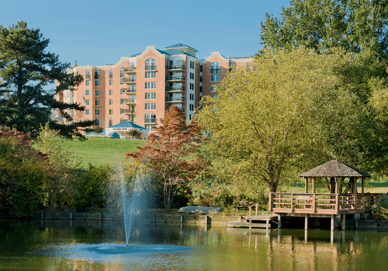 A large multi-story brick building in the background with balconies, surrounded by green trees and a grassy hill. In the foreground, there is a pond with a water fountain and a wooden gazebo on a dock extending over the water.