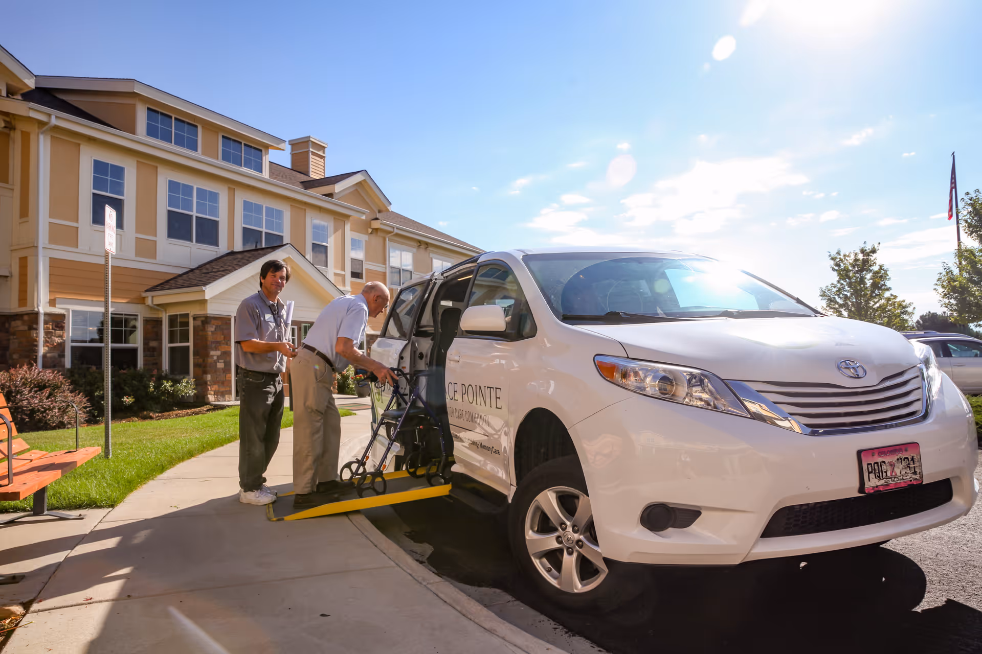 An elderly man using a walker is assisted by a caregiver as he boards a white van with a wheelchair lift outside a senior care community building on a sunny day.
