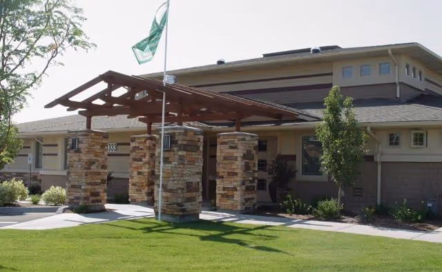 Front entrance of a single-story assisted living building with stone pillars, a wooden canopy, flagpole, and a manicured lawn.