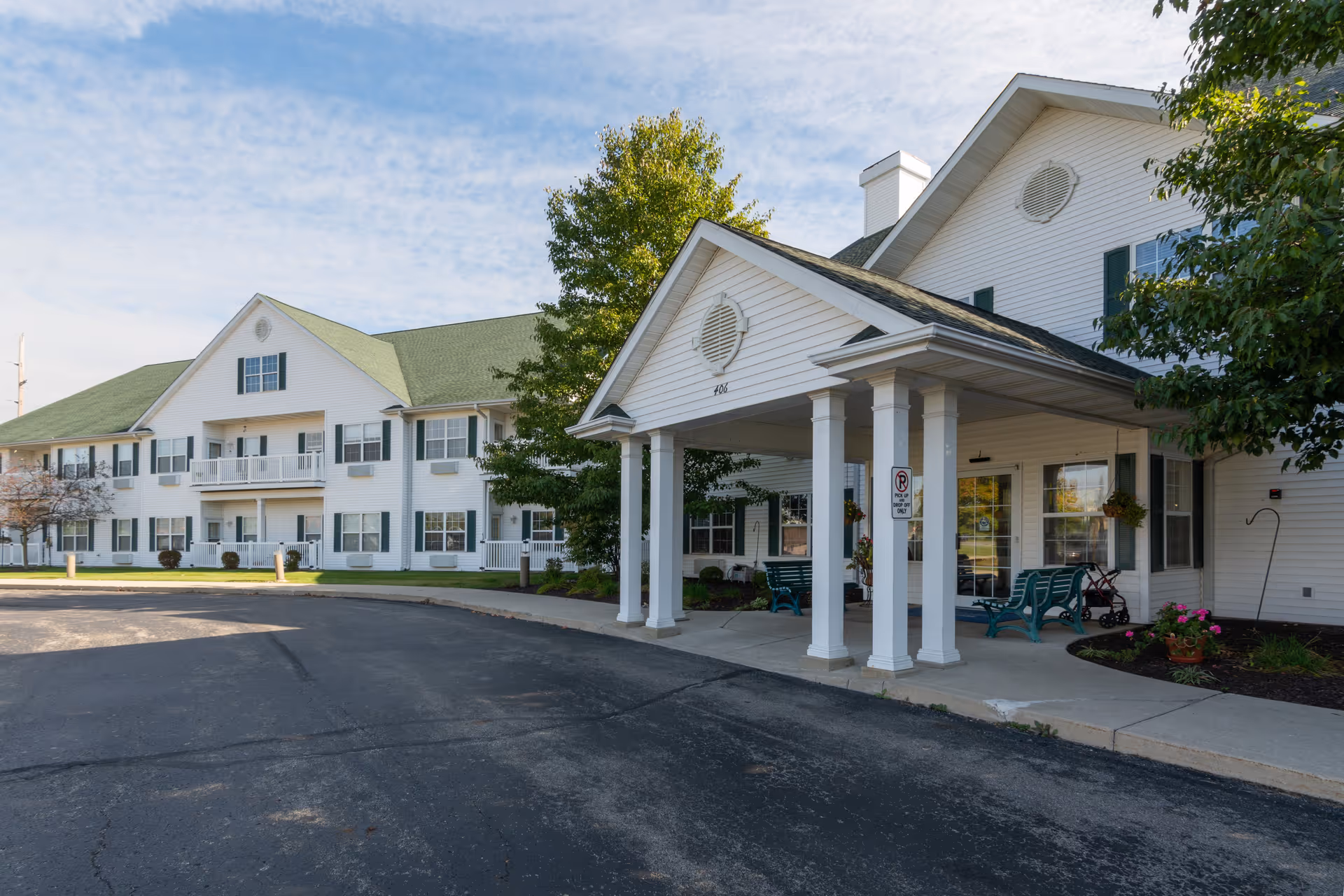Exterior view of Smith Farms Manor, a two-story white building with green shutters and a green roof. The entrance features a covered porch supported by white columns, with benches and potted plants nearby. Trees and a paved driveway are visible in front of the building under a partly cloudy sky.