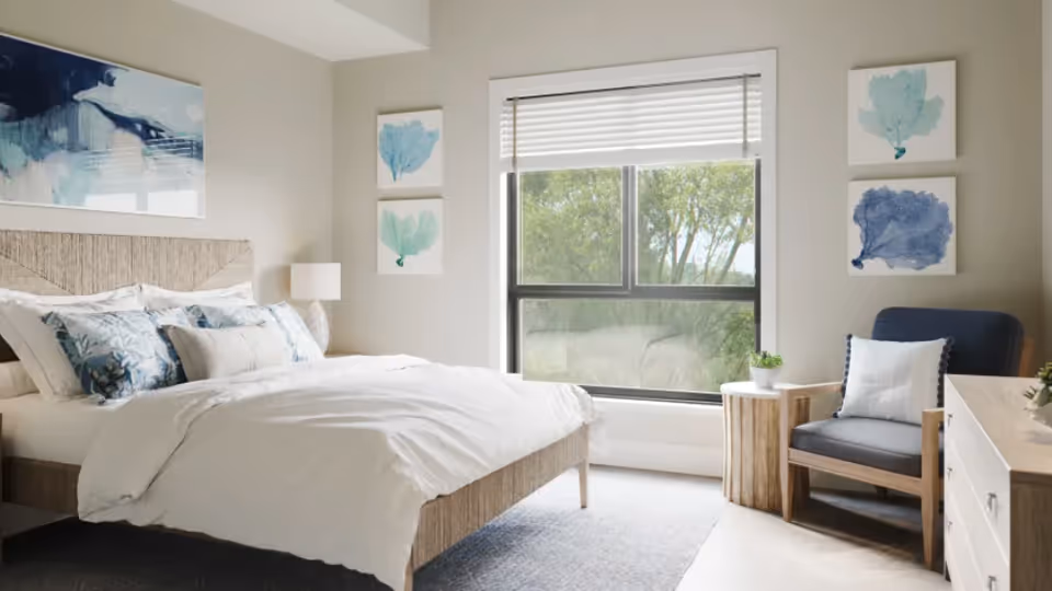 A bright and airy bedroom with a large bed featuring white and blue bedding, a wooden headboard, and a bedside lamp. The room has a large window with a view of green trees outside. There is a comfortable armchair with a white pillow, a small wooden side table with a plant, and a dresser. The walls are decorated with blue and teal abstract artwork.