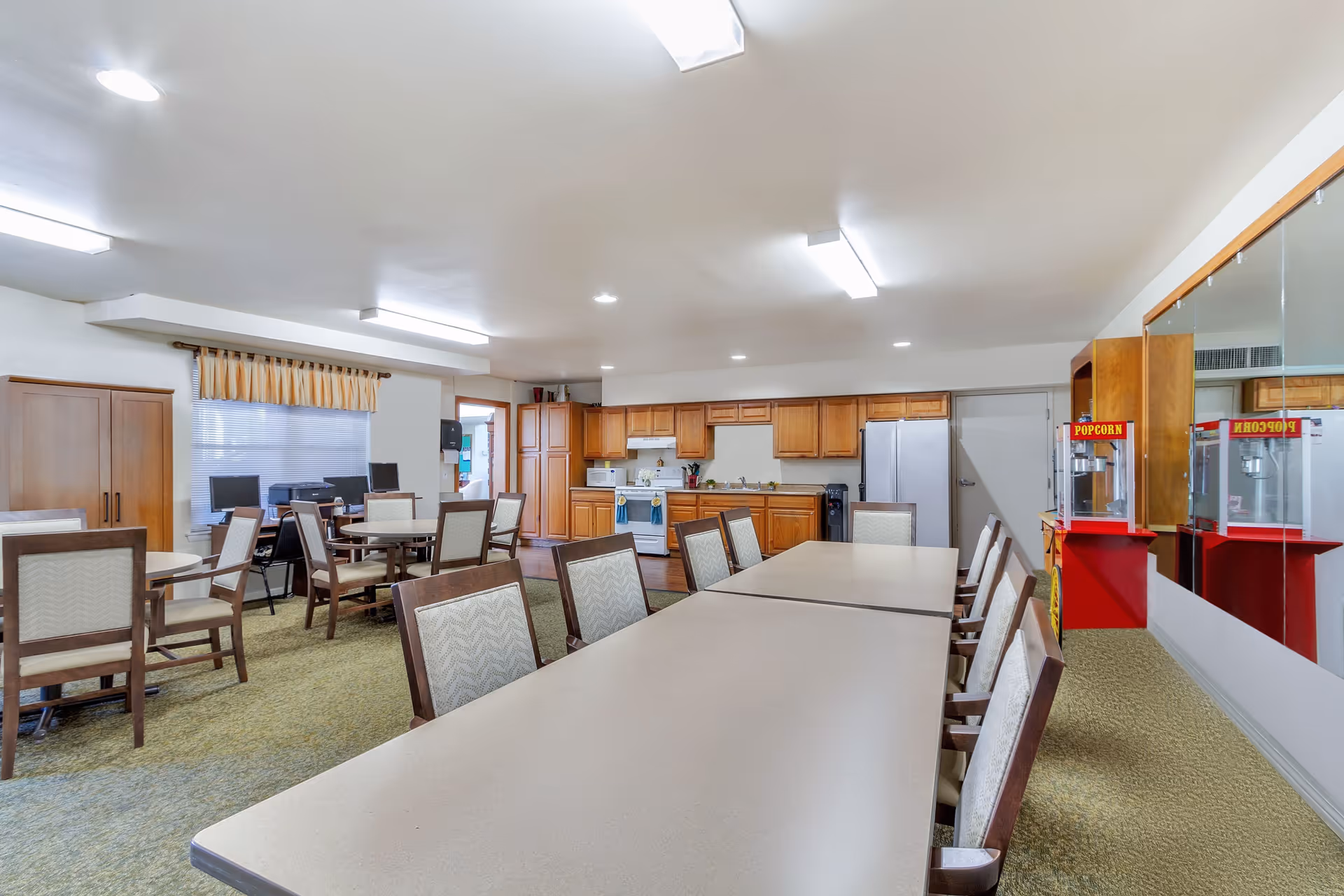 A spacious room with a long table and chairs in the foreground, smaller round tables with chairs in the background, a kitchen area with wooden cabinets, stove, microwave, and refrigerator along the back wall, and a popcorn machine near a mirrored wall on the right side.