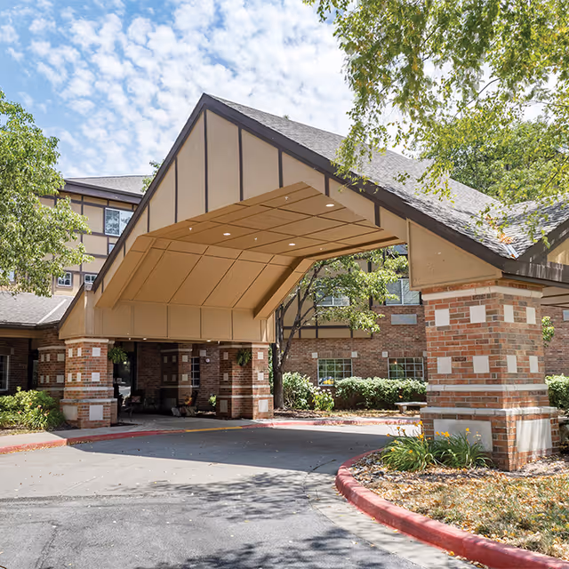 Entrance of a senior living facility with a covered drop-off area supported by brick pillars. The building has a mix of brick and beige paneling with dark trim. Trees and shrubs surround the entrance, and the sky is partly cloudy.