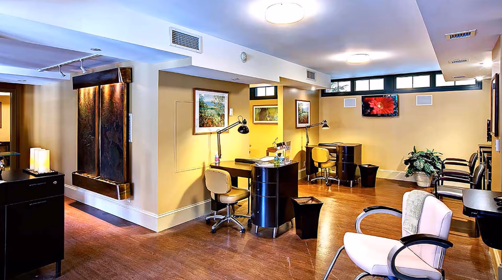 Interior view of a well-lit room with wooden flooring, featuring two black desks with yellow swivel chairs, desk lamps, and decorative wall art. There are additional chairs along the walls and a plant in the corner, suggesting a comfortable and functional common area.