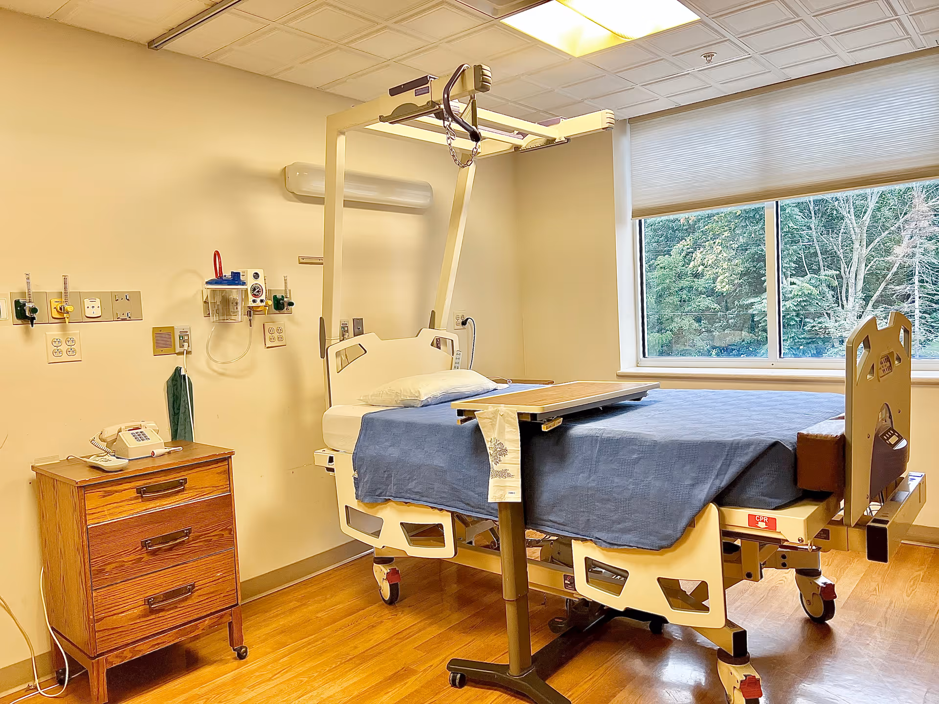 A hospital room with a medical bed covered with a blue blanket, a wooden bedside table with a telephone, medical equipment mounted on the wall, and a large window showing trees outside.