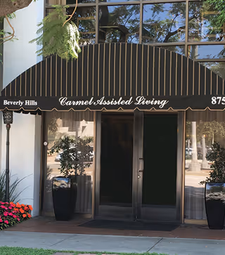 Entrance to a building with a black and beige striped awning that reads 'Beverly Hills Carmel Assisted Living'. There are two large black planters with green plants on either side of the glass double doors. Flowers and greenery are visible around the entrance.