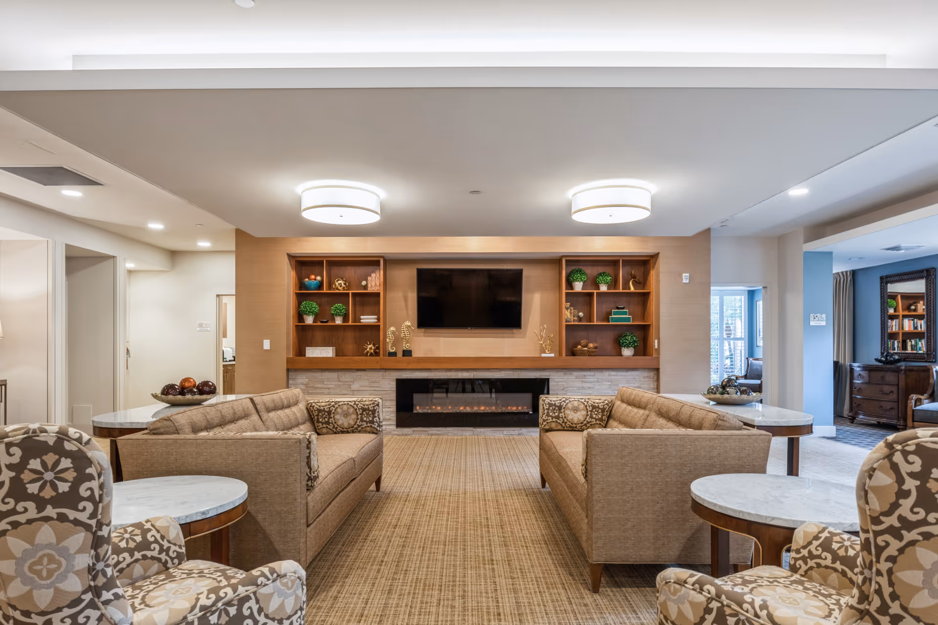 A modern living room with two beige sofas facing each other, a flat-screen TV mounted on a wooden wall unit with built-in shelves decorated with plants and ornaments, and a fireplace below the TV. There are patterned armchairs and round marble-top side tables in the foreground, with recessed lighting and ceiling fixtures illuminating the space.