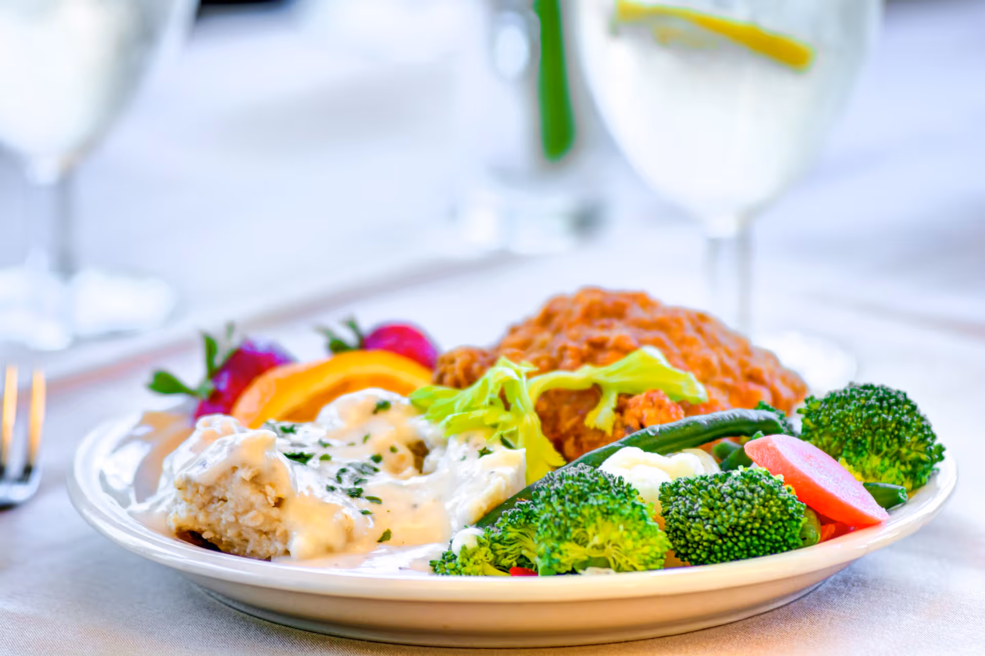 A plate of food featuring fried chicken, mashed potatoes with gravy, steamed broccoli, green beans, a slice of tomato, and a garnish of lettuce and fruit on a white tablecloth with glasses of water in the background.