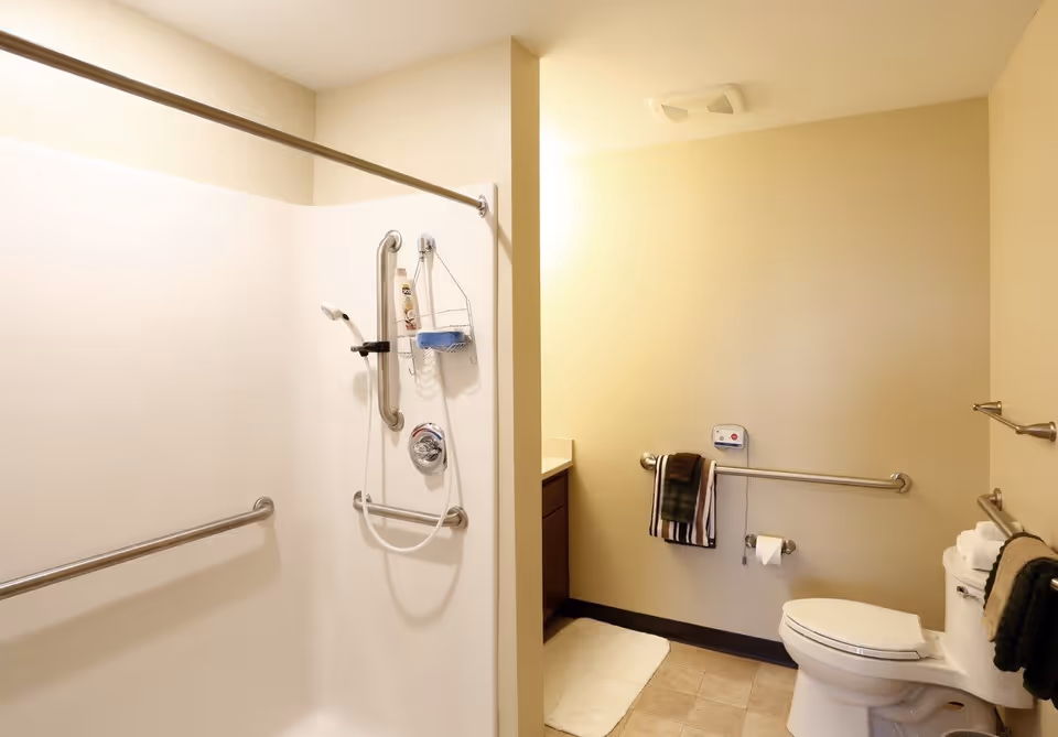 A clean and accessible bathroom featuring a walk-in shower with grab bars and a handheld showerhead. The bathroom also includes a toilet with grab bars on the walls, a towel rack with towels, a toilet paper holder, and a vanity with a countertop. The walls are painted beige and the floor is tiled.