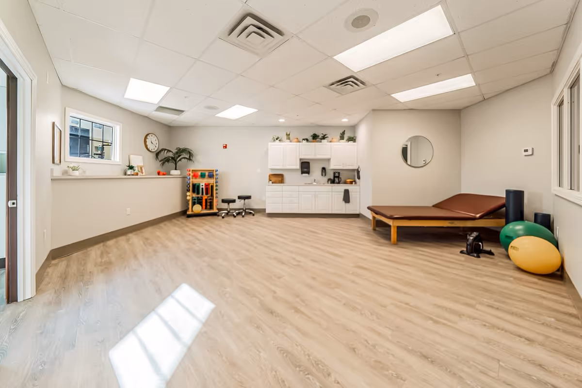 Bright multi-purpose therapy/exercise room with a treatment table, exercise balls, cabinets, and activity equipment on wood-look flooring.
