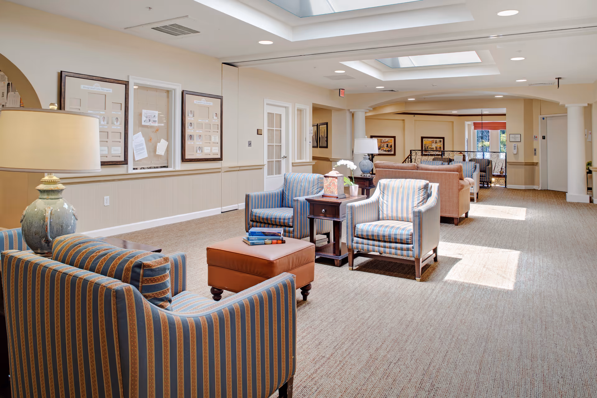 A bright and spacious senior living facility common area with striped armchairs, a brown leather ottoman, side tables with lamps and books, beige carpet, and skylights allowing natural light to fill the room. The walls are light-colored with framed pictures and a bulletin board.