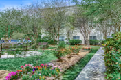 Landscaped garden path with flowering beds, trees, and a multi-story building facade in the background.