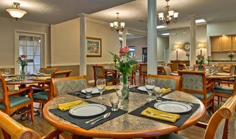 A well-lit dining room with multiple tables set for a meal, featuring plates, glasses, silverware, and yellow napkins. The room has wooden chairs with cushioned seats, decorative columns, chandeliers, and floral centerpieces on the tables. The background shows a kitchen area and framed artwork on the walls.