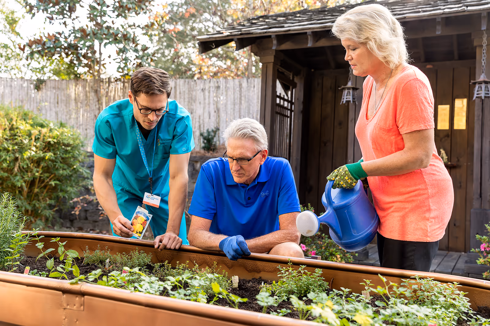 A man in a blue shirt and gloves and a woman in an orange shirt with gardening gloves are tending to a raised garden bed with various plants. A healthcare worker in teal scrubs is showing them a packet of seeds. They are outdoors in a garden area with trees and a wooden structure in the background.