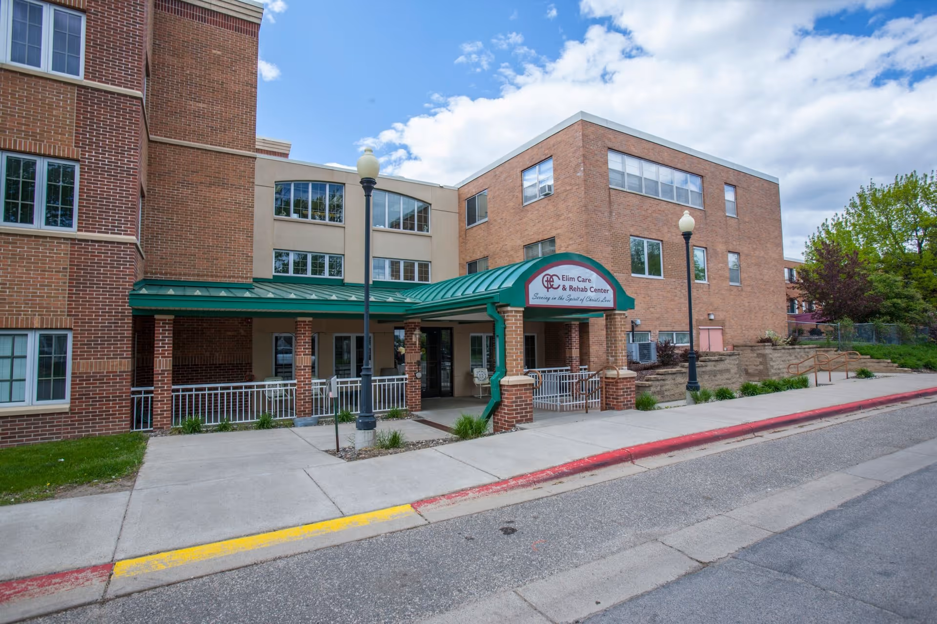 Front exterior of Elim Wellspring Health Care Center — a brick multi-story building with a green awning over the entrance and a sidewalk.