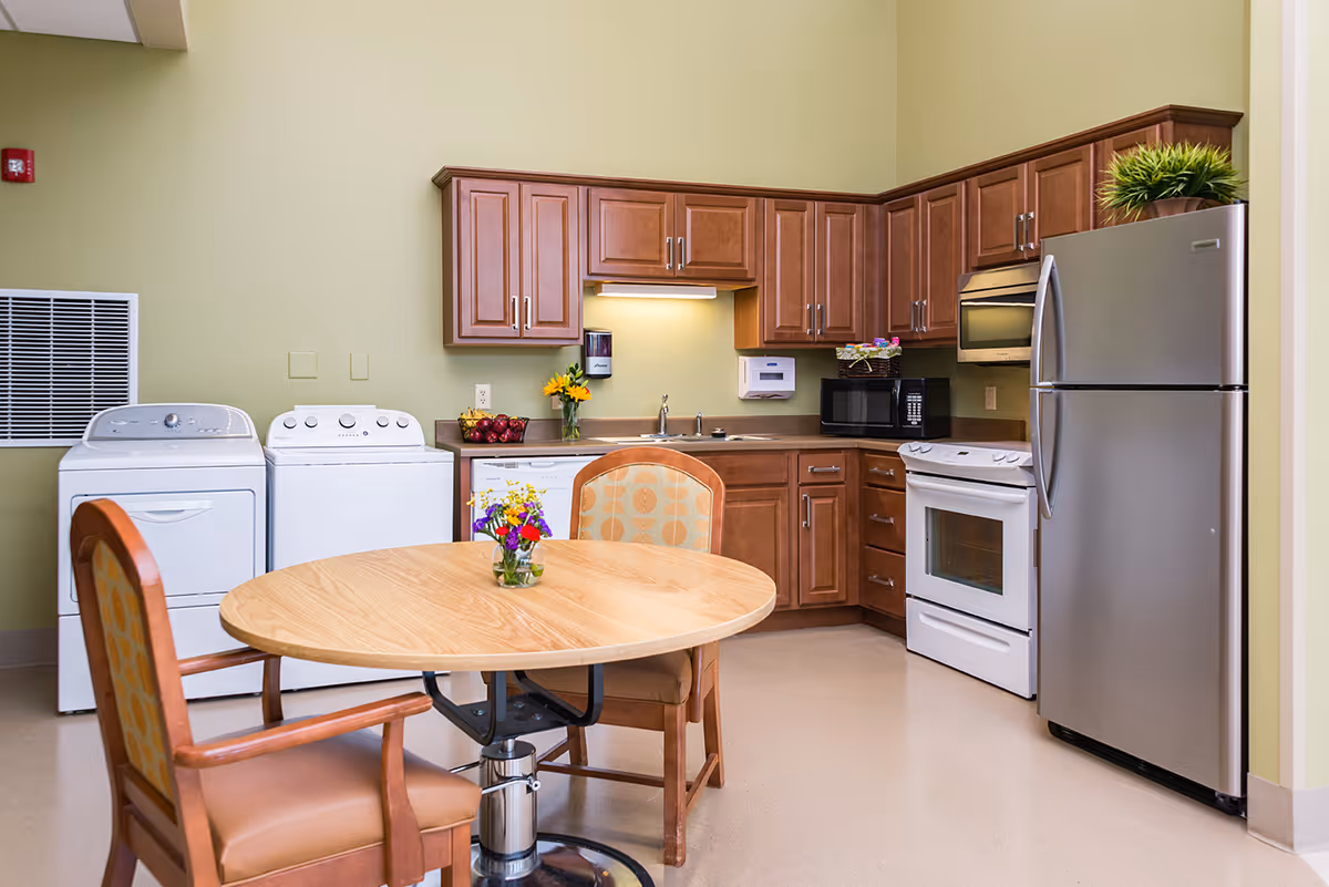 A clean and well-lit kitchen area with wooden cabinets, a stainless steel refrigerator, a white stove, microwave, dishwasher, and a washer and dryer set against a light green wall. In the foreground, there is a round wooden table with two cushioned chairs and a small vase of colorful flowers on the table.