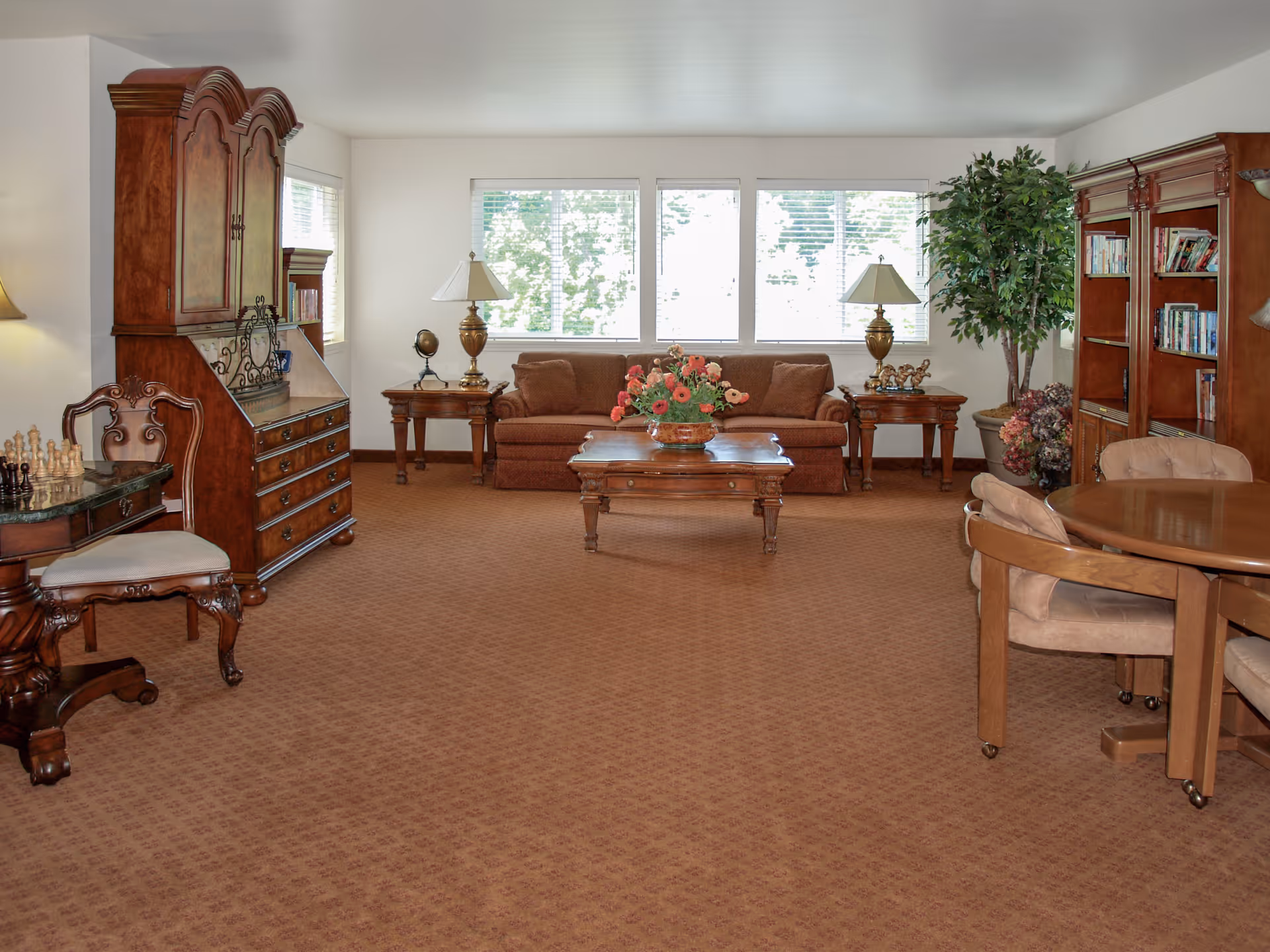 A spacious living room with brown carpet and wooden furniture including a sofa, coffee table with a flower arrangement, side tables with lamps, a large wooden cabinet, a bookshelf filled with books, a round table with chairs, and a chess set on a small table. Large windows in the background let in natural light, and there is a potted plant in the corner.