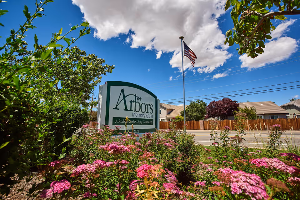 Arbors Memory Care sign in a landscaped front with pink flowers, an American flag, and houses in the background.
