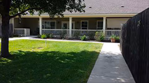 Front porch of a single-story building with a covered veranda, pathway, lawn, tree, and a fenced side yard.