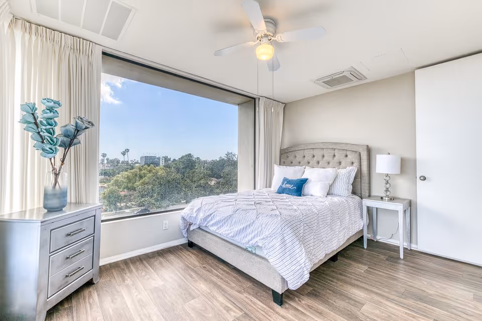 A bright bedroom with a large window showing a view of trees and buildings outside. The room features a bed with a tufted headboard, white and striped bedding, and a blue pillow with the word 'home' on it. There is a silver nightstand with a lamp on one side of the bed and a silver dresser with a vase of blue flowers on the other side. The floor is wooden, and a ceiling fan with a light is mounted on the white ceiling.