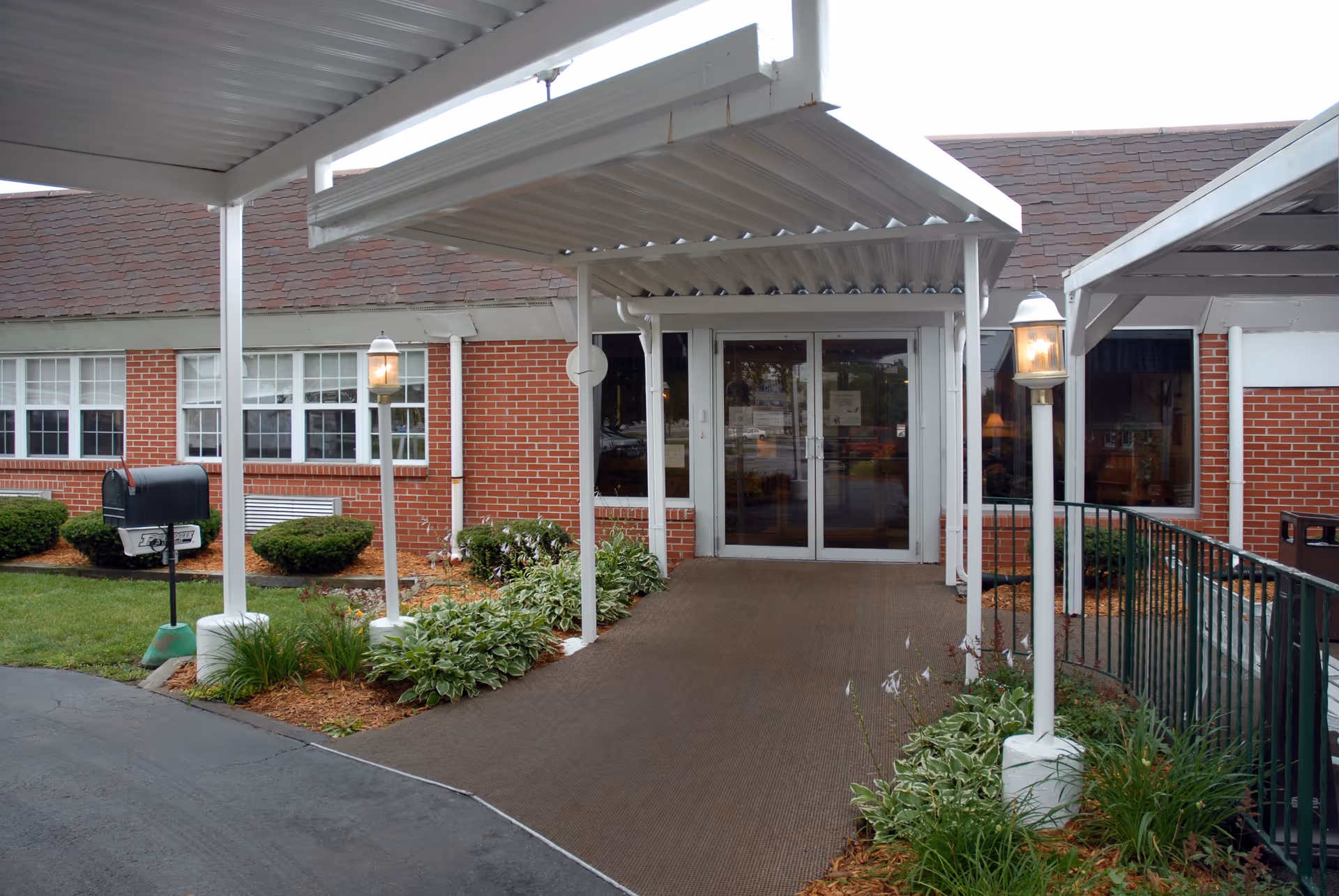 Covered entrance walkway leading to glass double doors of a brick building with lamps and landscaping.