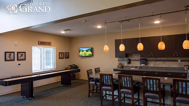 Interior view of a common area at The Oxford Grand featuring a shuffleboard table, a high counter with six chairs, pendant lights hanging above the counter, cabinets, and a wall-mounted TV. The room has beige walls, carpeted floor, and framed artwork on the walls.