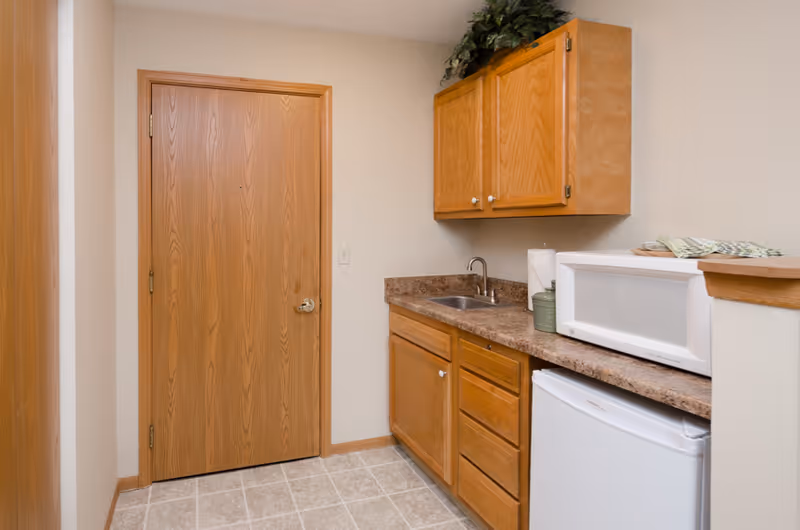 Small kitchenette area with wooden cabinets, a countertop with a sink, a white microwave, and a mini refrigerator. There is a wooden door on the left side and beige tiled flooring.