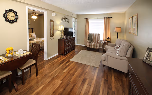 A furnished living room with hardwood floors, a sofa and armchair, TV on a dresser, a small dining table, and a doorway leading to a bedroom.
