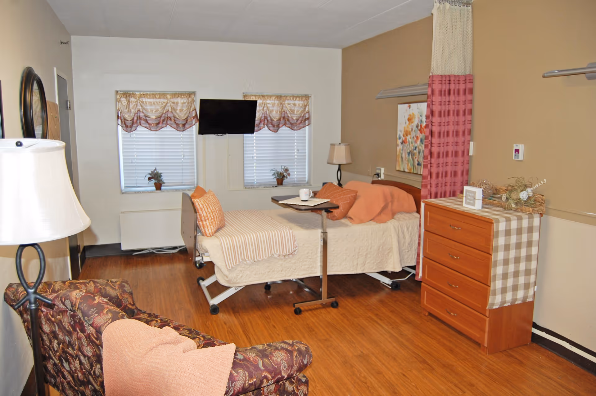 A nursing facility room with a hospital bed covered in beige bedding and orange pillows, a small overbed table with a cup and papers, a wooden dresser with a checkered cloth and decorative items, a patterned armchair with an orange pillow, two windows with blinds and valances, a wall-mounted TV, a floor lamp, and a privacy curtain partially drawn.