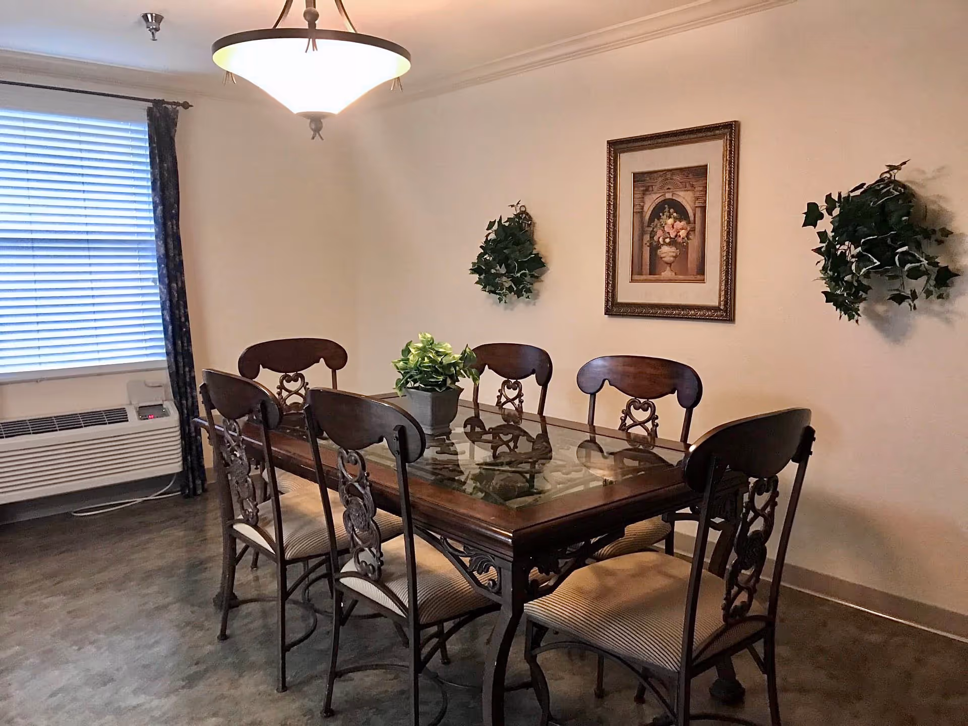 A dining room with a rectangular glass-top wooden table surrounded by six ornate wooden chairs with cushioned seats. A potted plant is placed in the center of the table. The room has a window with closed blinds and dark curtains, a wall-mounted air conditioning unit below the window, a ceiling light fixture, two wall-mounted green plants, and a framed floral artwork on the wall.