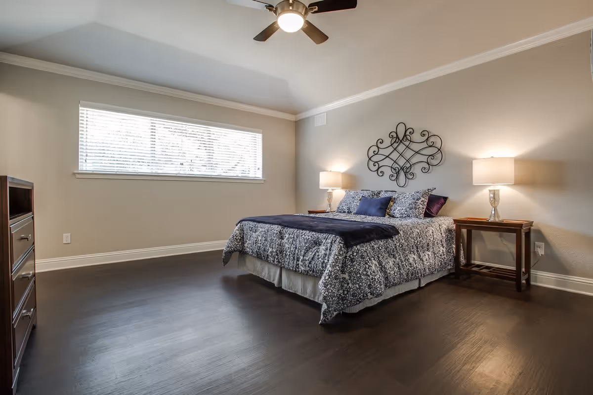 A bedroom with a large bed covered in a patterned blue and white comforter, two bedside tables with lamps, a decorative metal wall art above the bed, a ceiling fan with light, a wide horizontal window with blinds, and dark wood flooring.